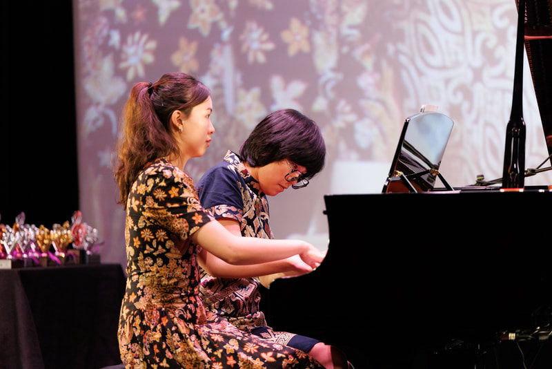 Two women sit at a grand piano performing on stage, with a colorful floral background projection and a display of trophies on a table nearby.