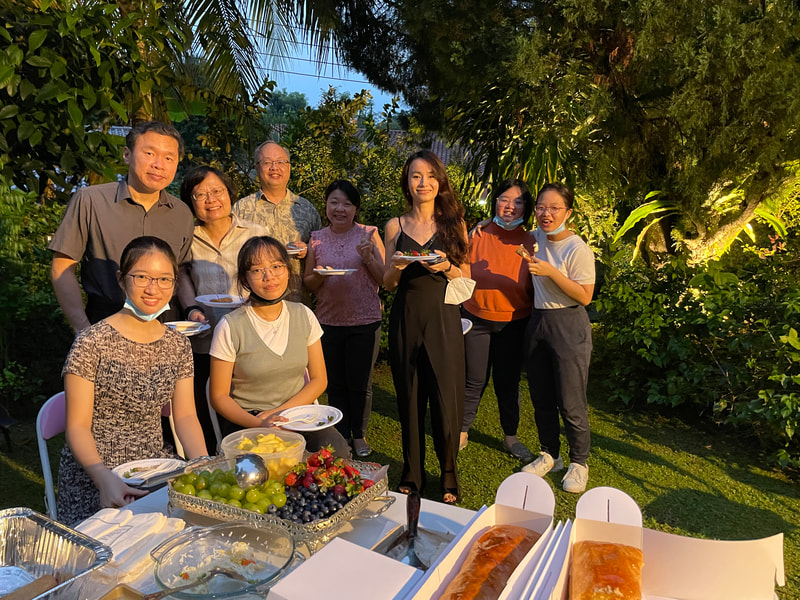 A group of nine people, mostly women, standing and sitting outdoors in a garden during sunset, celebrating with a table full of desserts and fruits, smiling at the camera.