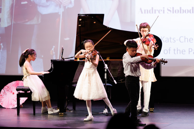 Children performing a musical recital on stage, with a girl playing piano, two girls and a boy playing violins.