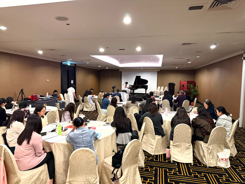 A conference room filled with attendees sitting at round tables, facing a stage with a grand piano and a white projection screen, in a professional setting.