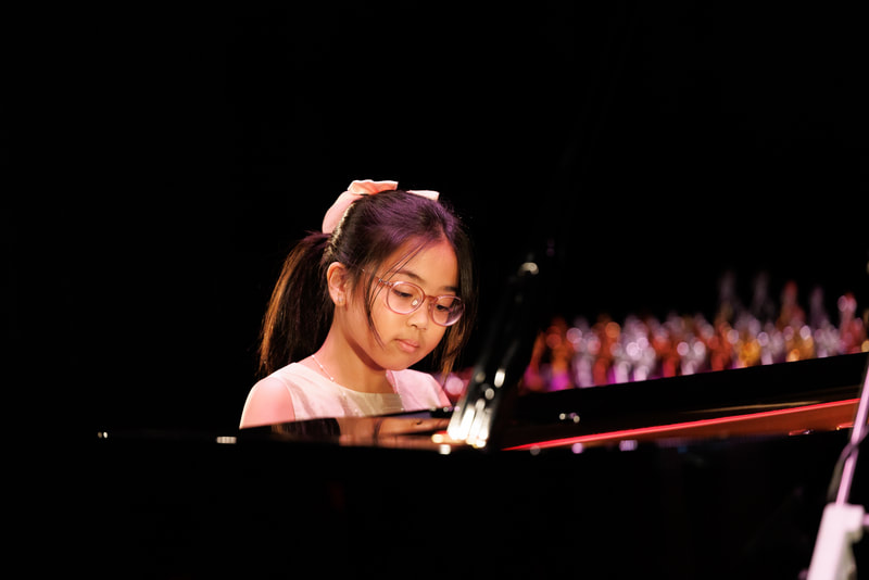 A young girl with glasses playing a grand piano on stage during a performance.