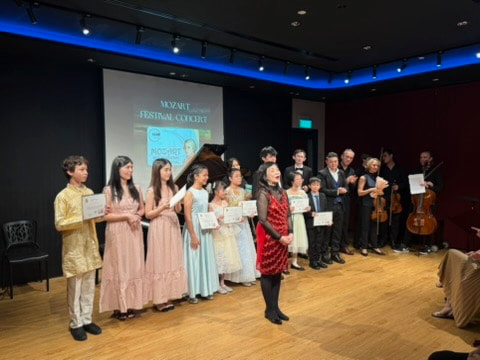 Young children and adults standing on stage during a concert, holding certificates, with a piano and a large screen displaying text in the background.