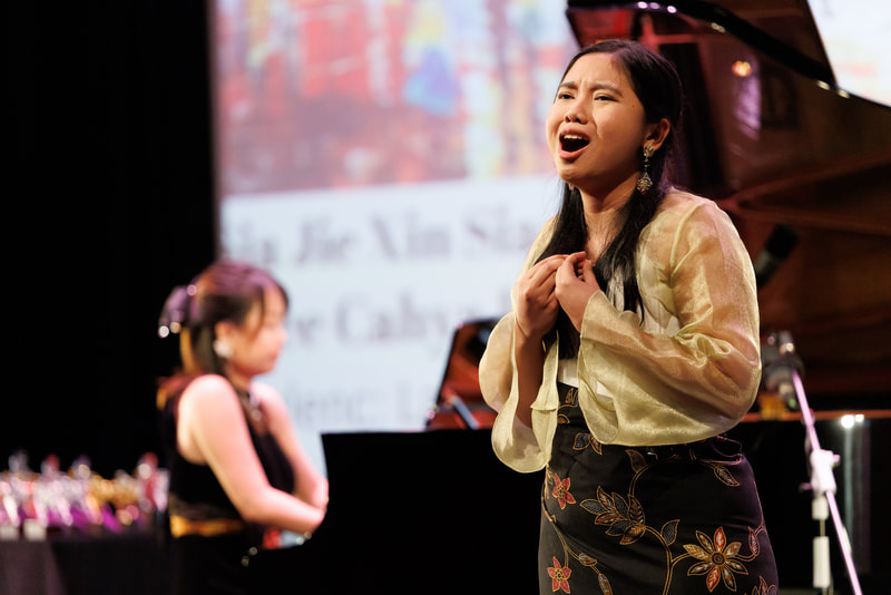 A woman singing passionately on stage with a pianist in the background, during a musical performance.