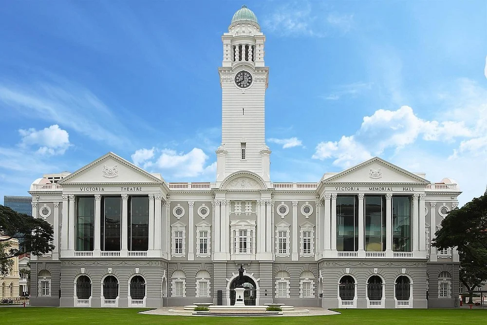 White historic building with a central clock tower, labeled as Victoria Theatre and Victoria Memorial Hall, under a blue sky with some clouds.