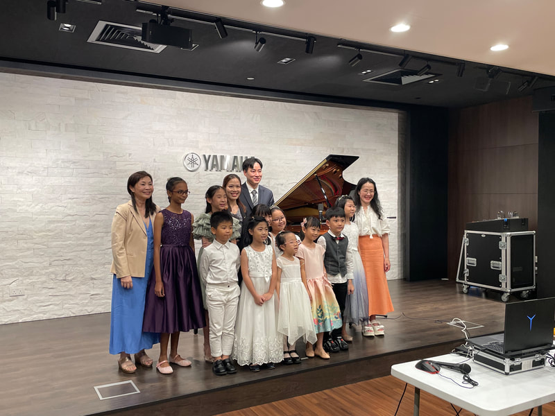 Group of children and adults posing on a stage with a grand piano in the background at Yamaha, some children are holding certificates or awards.