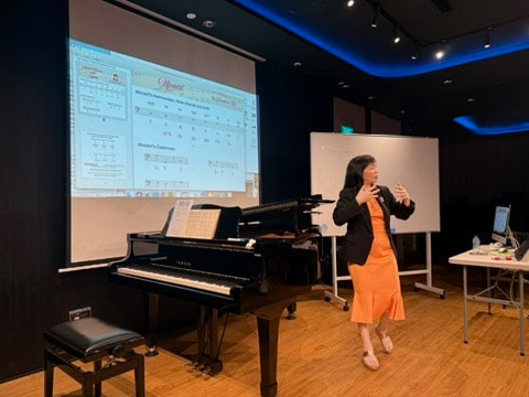 A woman standing and presenting in a classroom or lecture hall, with a large projector screen displaying a spreadsheet or software interface, a black grand piano, a whiteboard, and desks with computer equipment.