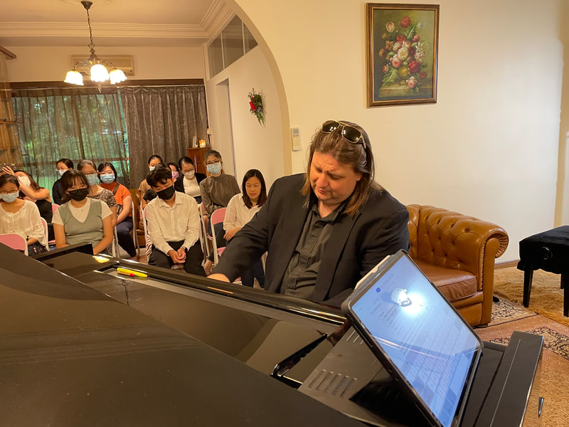 Woman playing piano in a living room while a group of children and adults watch, all wearing masks.