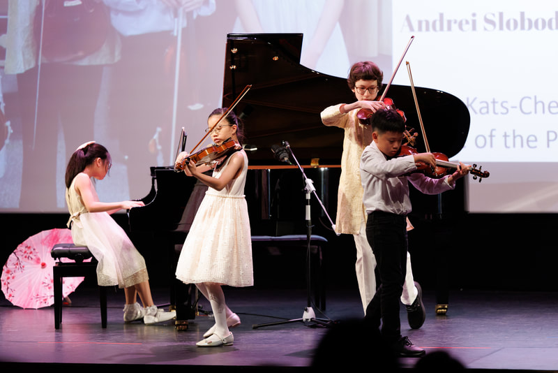 Children and an adult playing violins on stage, with a grand piano and a projection screen in the background.