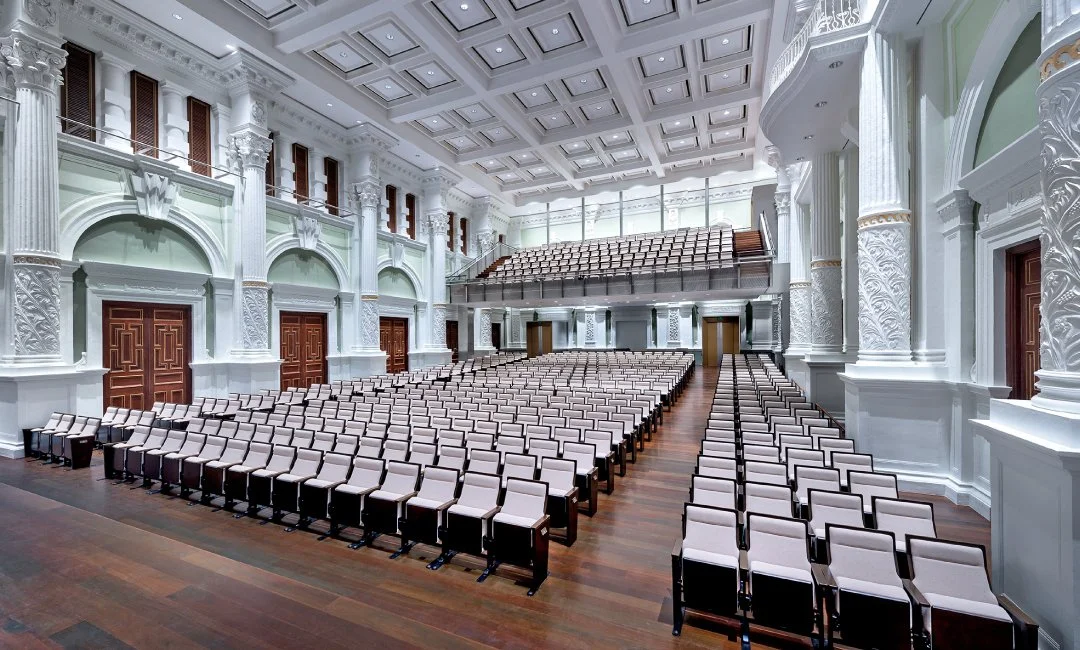 Large, ornate auditorium with rows of empty white chairs, high ceilings with intricate molding, and decorative architectural details.