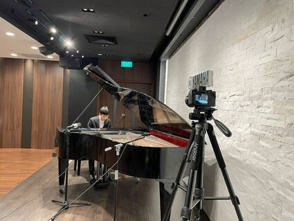 Person playing grand piano in recording studio with camera on tripod.