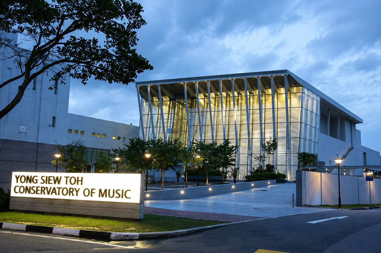 The Yong Siew Toh Conservatory of Music building at dusk, featuring modern architecture with glass and metal design, illuminated from within, with a sign in the foreground reading "Yong Siew Toh Conservatory of Music," surrounded by trees and streetlights.