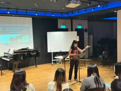 A woman presenting on stage in front of seated audience, with a whiteboard, a grand piano, and a projector screen displaying a presentation slide in a room with blue lighting.