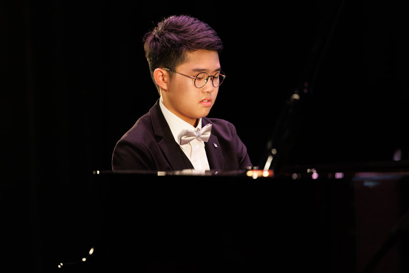 Young man wearing glasses and a tuxedo with a bow tie playing a grand piano on a dimly lit stage.