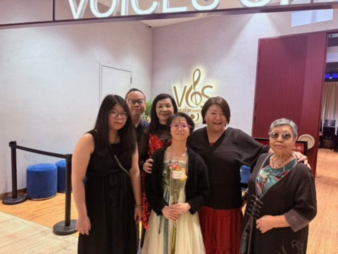 Five people standing together in an indoor setting with a sign that says 'Voices' background, some holding bouquets, smiling, dressed in semi-formal to formal clothing.