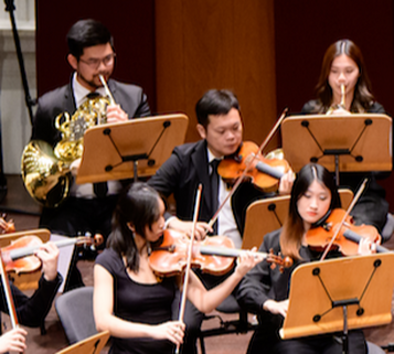 Orchestra musicians performing on stage, playing violins and a saxophone, with music stands in front of them.