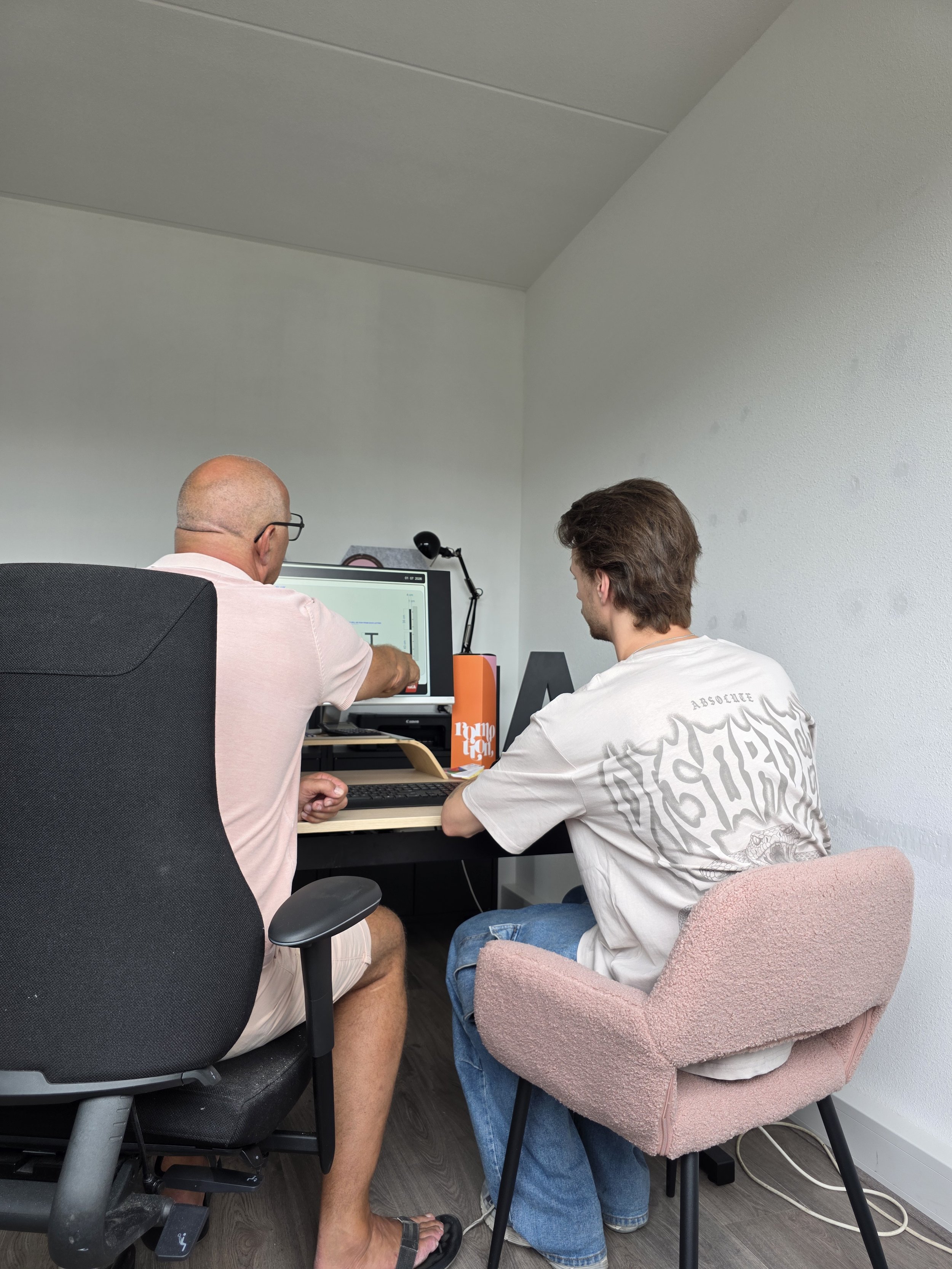 Twee mannen werken samen aan een computer in een kantoor. Ze are zittend, één met een roze polo en bril, de ander in een witte T-shirt en jeans. Ze bekijken het scherm, met één op een bureaublad en de ander op een pinkelstoel. Er bevinden zich boeken en decoraties op de tafel.