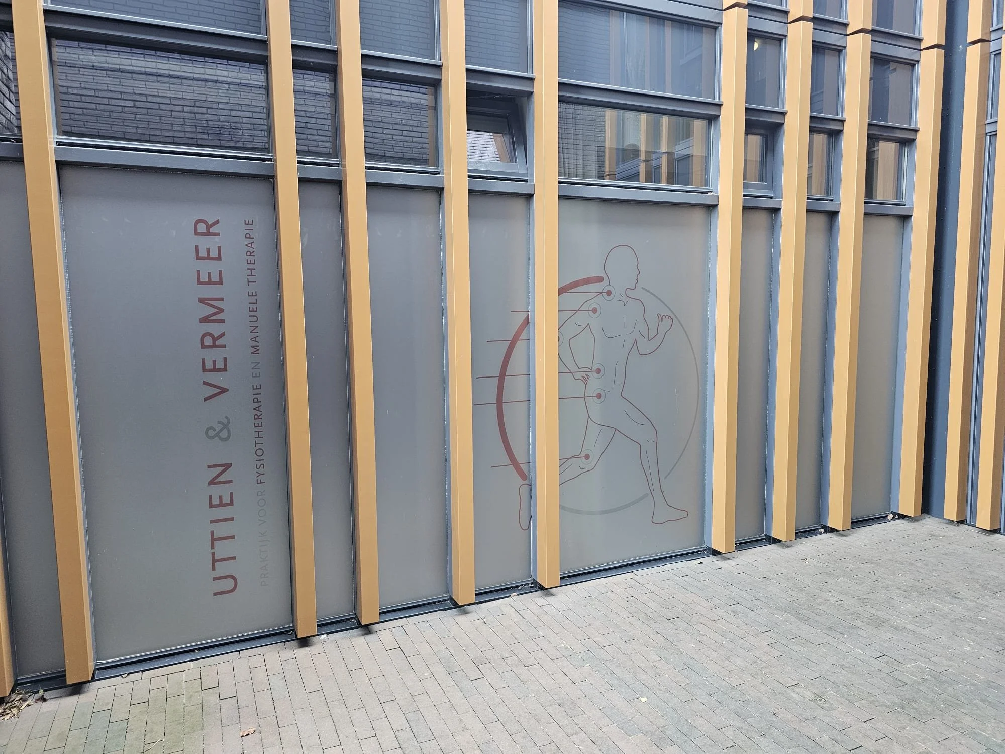 De afbeelding toont een modern gebouw met grote ramen en verticale gele en grijze panelen. Op een venster is de tekst zichtbaar: 'VOORNE MER' en onderaan 'THERAPIE'.