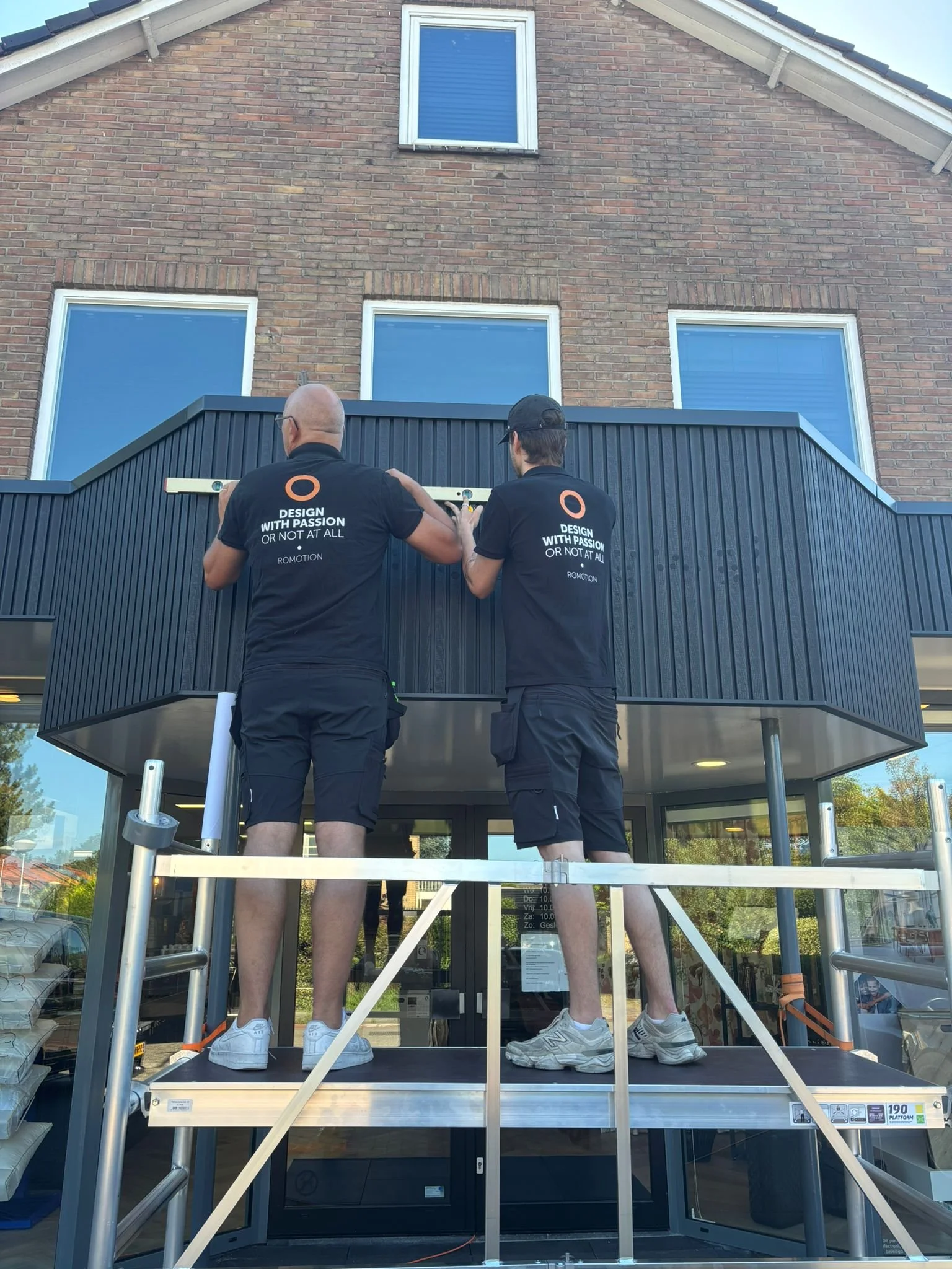 Twee mannen staan op een hoog werkplatform voor een gebouw met een bakstenen gevel, bezig met het installeren of meten van het gebouw. Ze dragen zwarte shirts met de tekst 'Design with Passion or not at all'.
