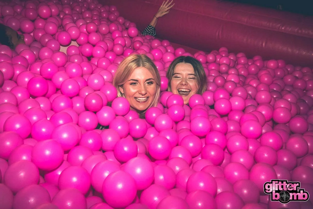 Two women peering over the edge of a bright pink ball pit