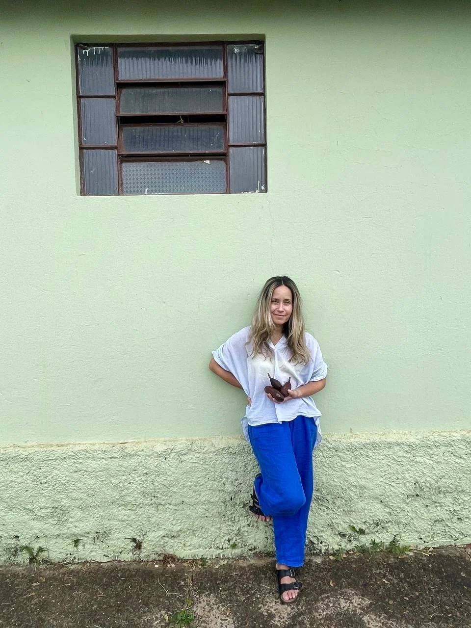 A woman with long blonde hair dressed in a white shirt and blue pants stands leaning against a light green wall outside a building. She is holding a dark-colored butterfly in her hands and barefooted.