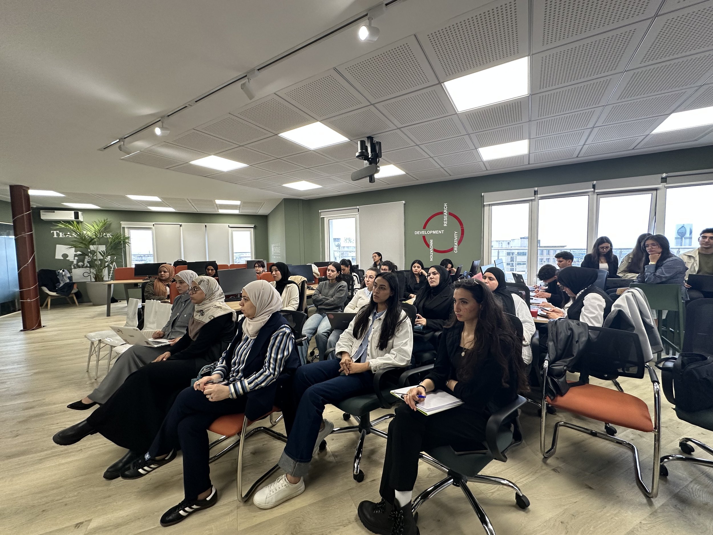 Une salle de conférence ou de formation remplie de personnes assises en rangées, écoutant attentivement. La pièce a de grandes fenêtres laissant entrer de la lumière naturelle, un mur vert avec des mots clés tels que "Innovation, Recherche, Créativité, Développement". Il y a aussi des plantes vertes et une ambiance professionnelle.