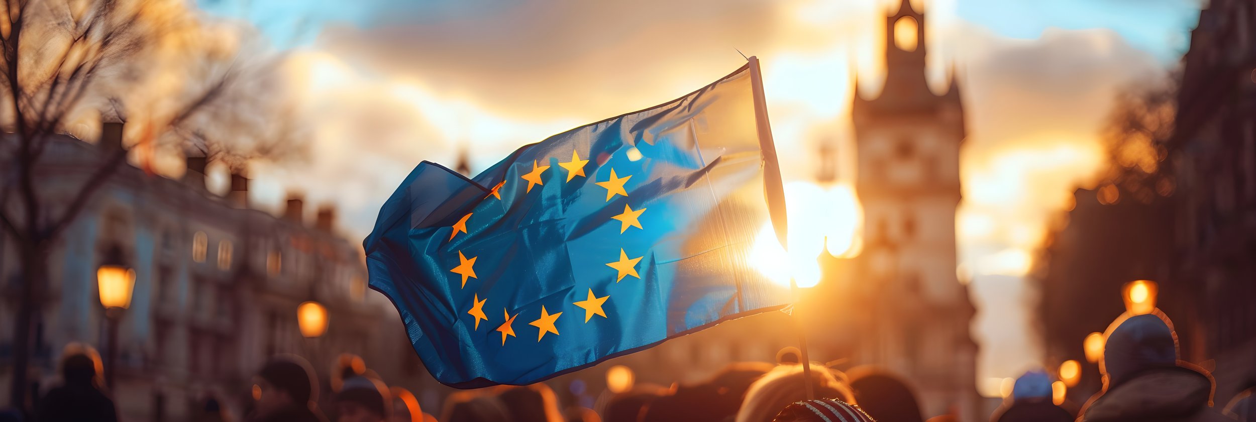 Drapeau de l'Union européenne flotte lors d'un coucher de soleil dans une ville, avec des silhouettes de personnes en arrière-plan.