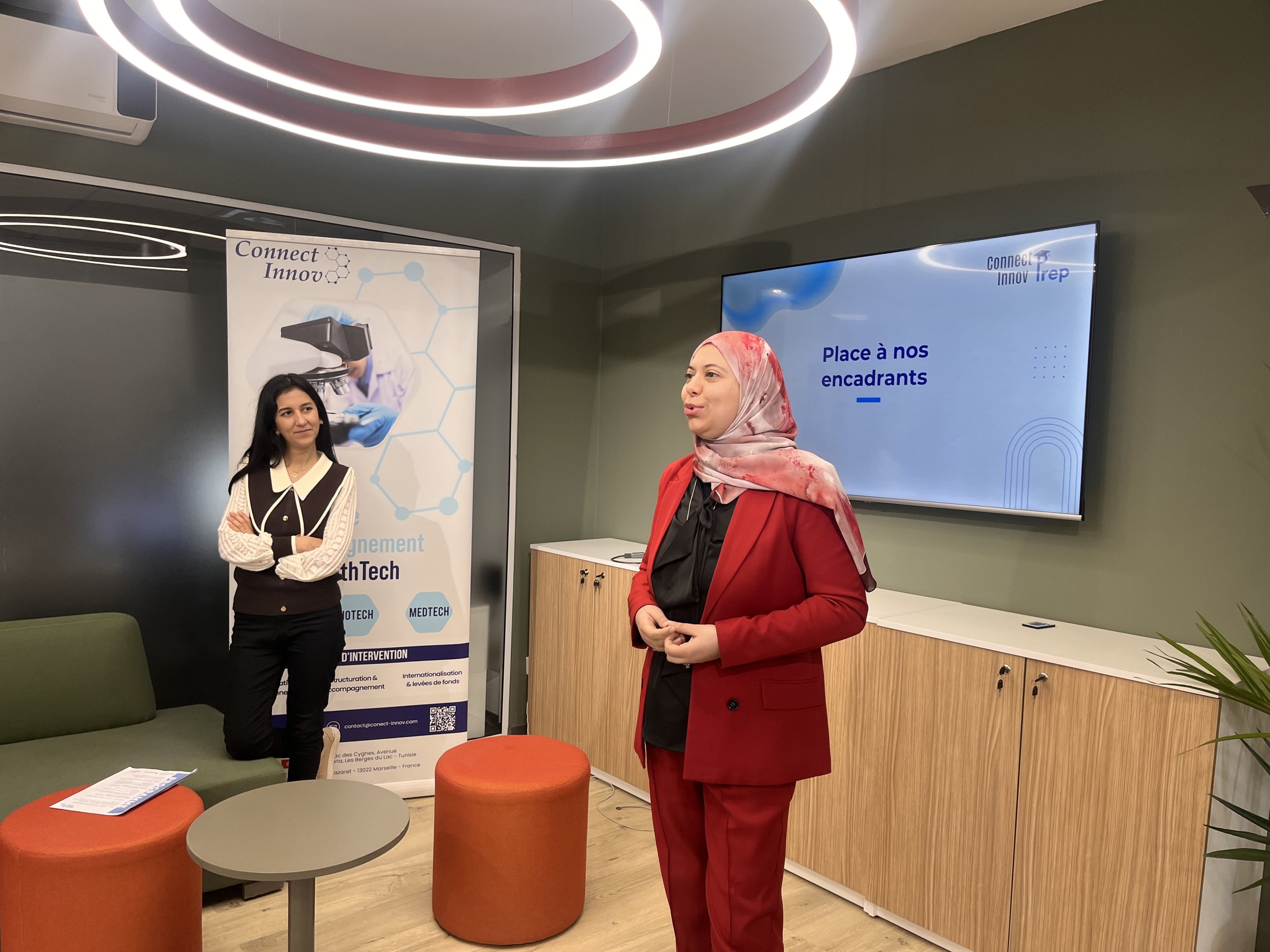 Deux femmes discutent dans une salle de réunion moderne, avec un écran numérique affichant le logo "Connect Innov" et la phrase "Place à nos encadrants". Une affiche promotionnelle se trouve à côté, avec du texte et un graphique représentant la connectivité technologique.