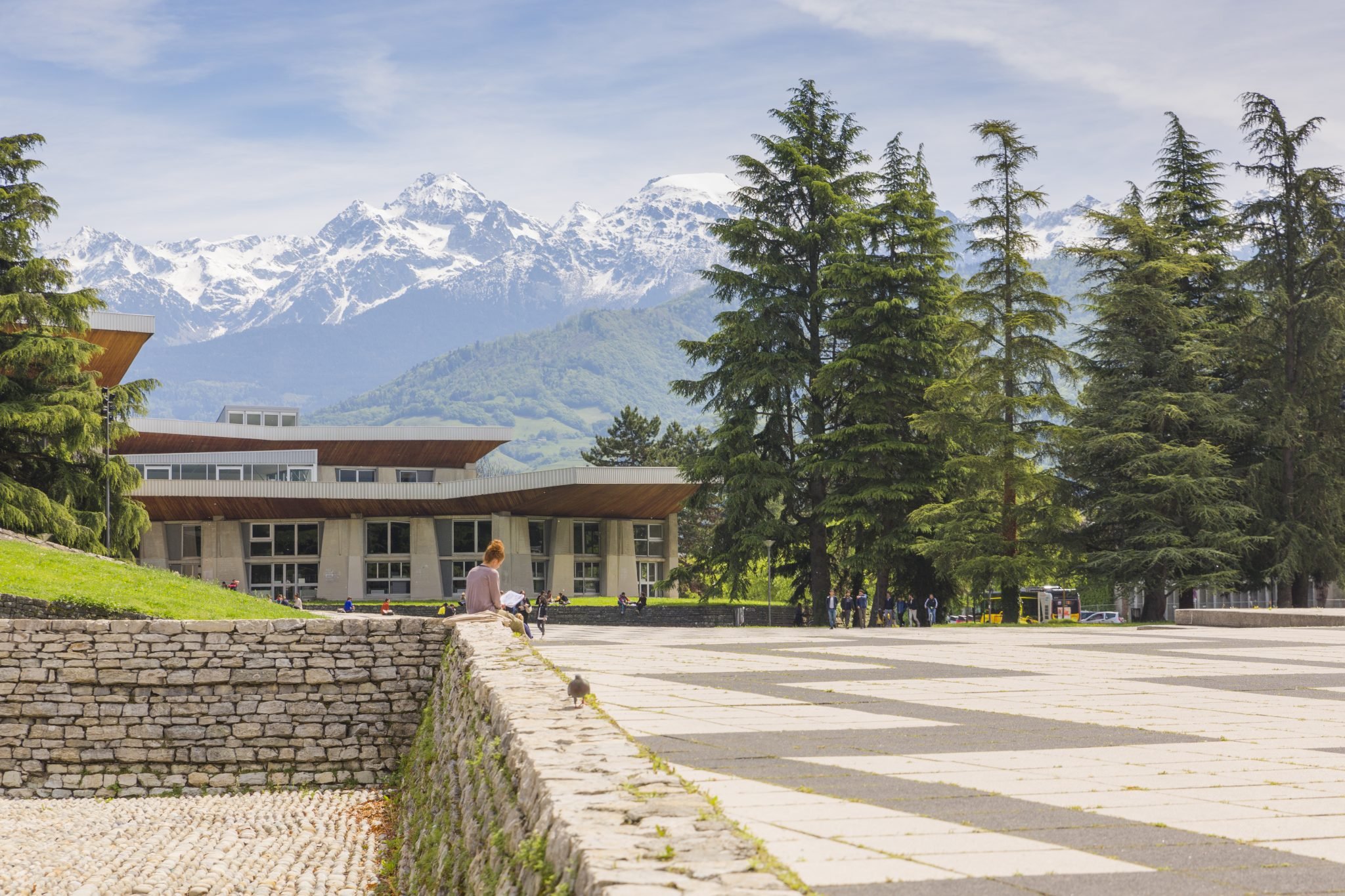 Un paysage avec des montagnes enneigées en arrière-plan, des arbres verts en premier plan, et un bâtiment moderne avec une terrasse en terrasse. Il y a aussi des personnes assises et marchant dans l'espace ouvert.