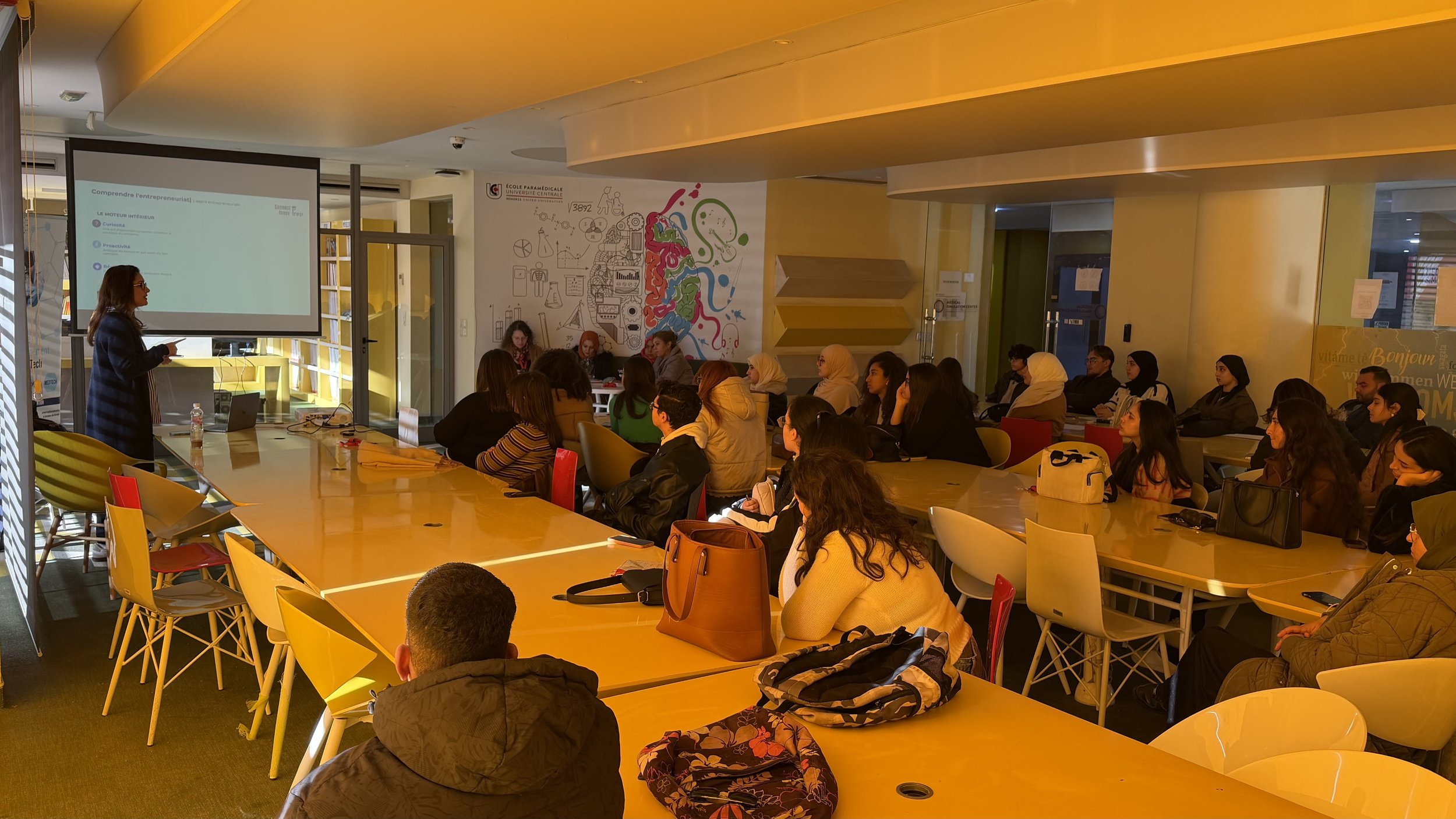 Une salle de classe ou de formation avec de nombreux élèves assis à une grande table, écoutant une femme qui fait une présentation devant un écran. Les murs sont jaunes avec des décorations éducatives.