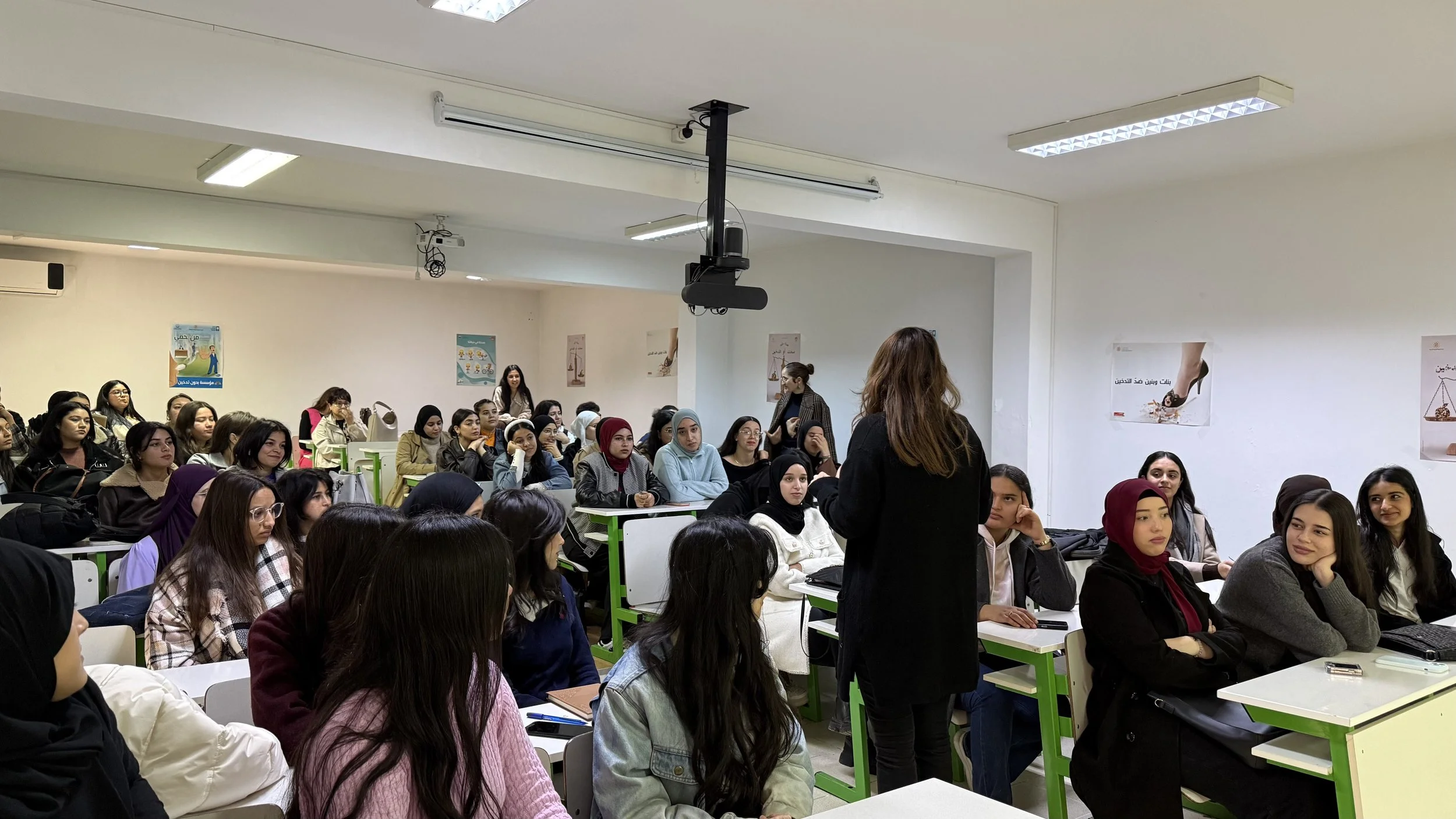 Une salle de classe remplie de jeunes femmes assises, quelques-unes portant des hijabs, écoutant une femme debout qui parle à l'avant.