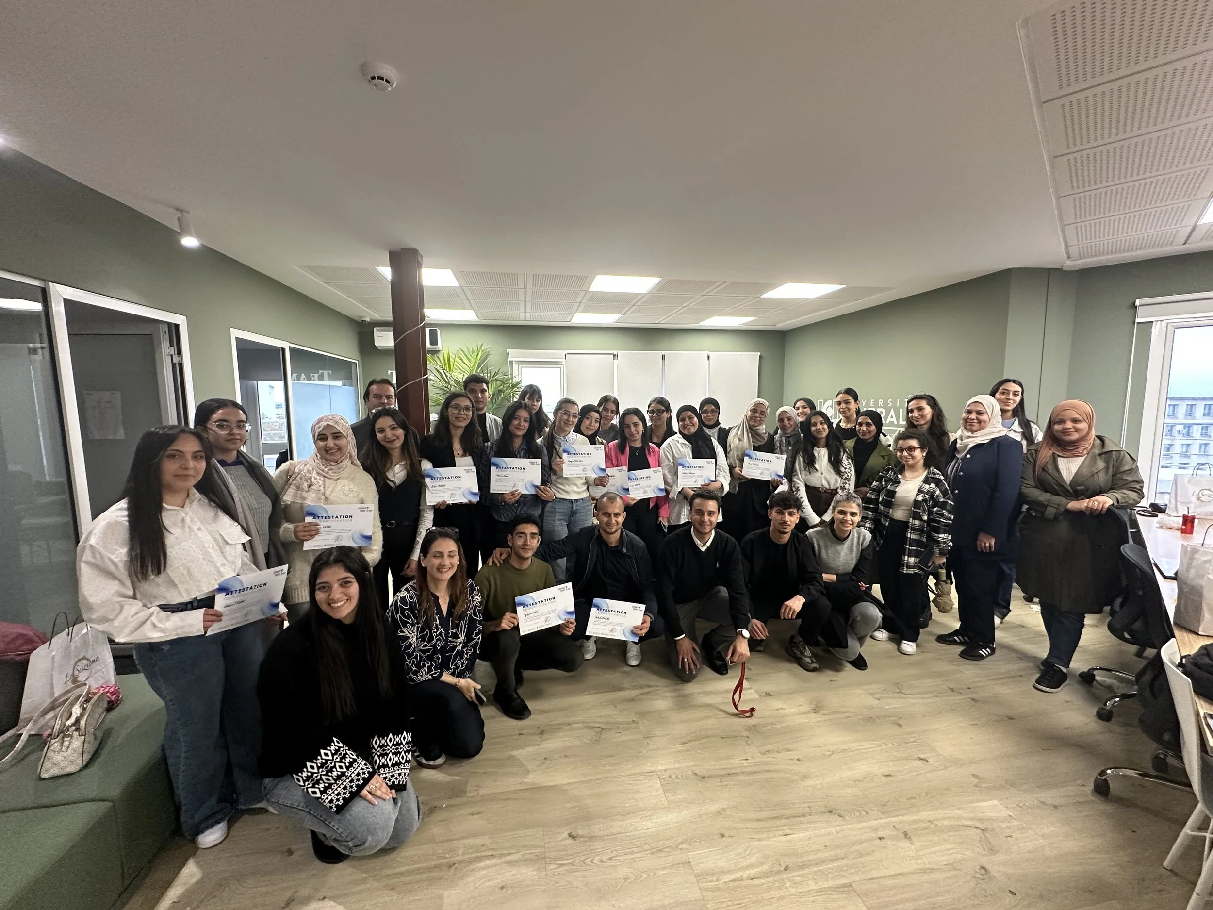 Groupe de personnes posant pour une photo, la plupart tenant des certificats, dans une pièce avec des fenêtres et un mur vert.