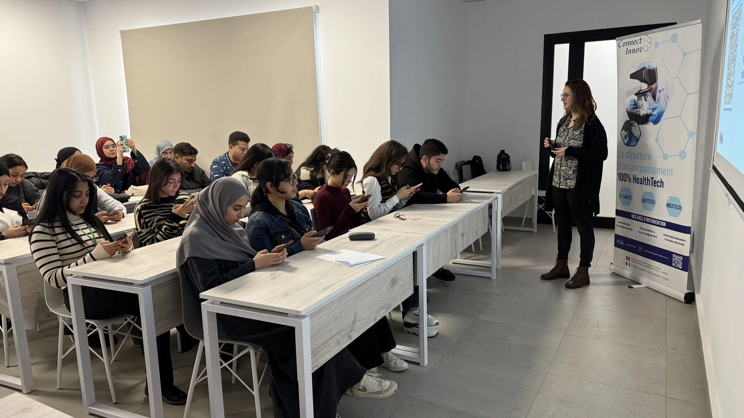 Une femme donne une présentation à un groupe d'étudiants assis à une table longue, utilisant leur téléphone, dans une salle de classe ou une salle de formation. Il y a un panneau d'information sur la droite avec des images de technologie médicale et des textes en français.