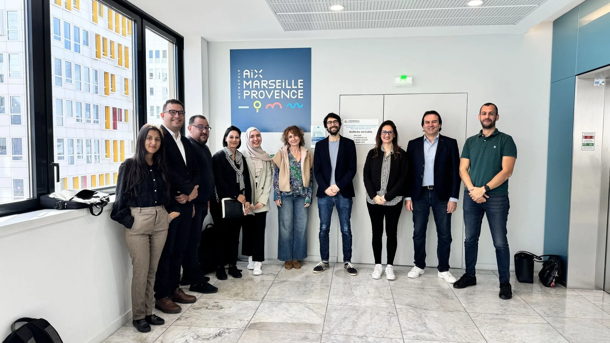 Groupe de personnes posant pour une photo devant une grande affiche bleue avec les noms Aix, Marseille, Provence, Métropole, dans un bureau ou une salle de réunion.