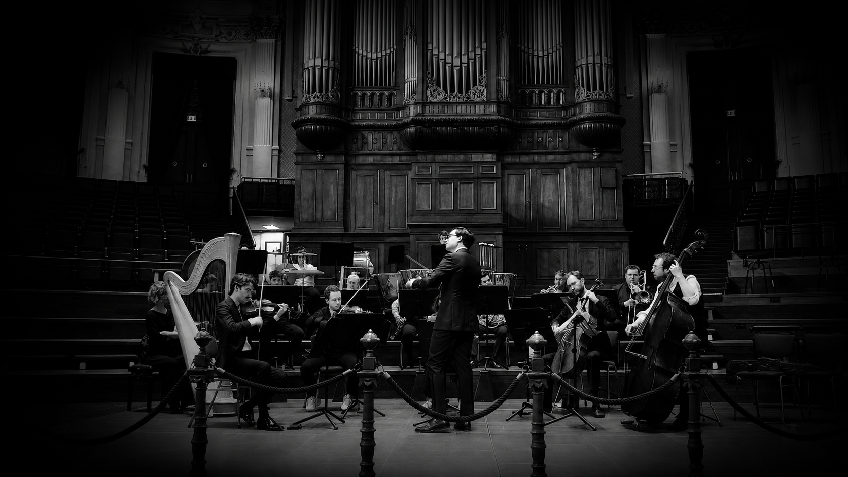 An orchestra performing in a grand hall with wooden paneling and an organ in the background. The conductor is standing in front of the musicians, who are playing various instruments including a harp, violins, and a double bass. The scene is black and white.