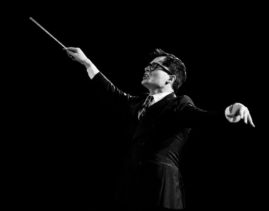 A man in a suit and glasses conducting an orchestra with a baton on a dark stage.