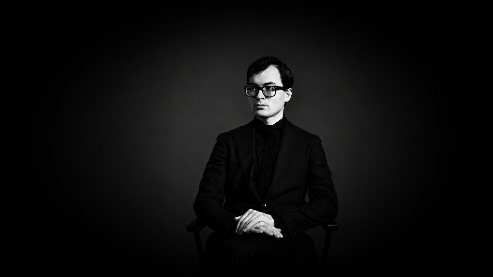 Black and white portrait of a young man in a suit and glasses sitting with hands clasped, looking to the side against a dark background.