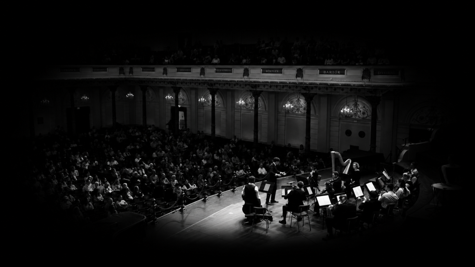 Black and white photo of an orchestral concert in a grand concert hall with an audience in the background.