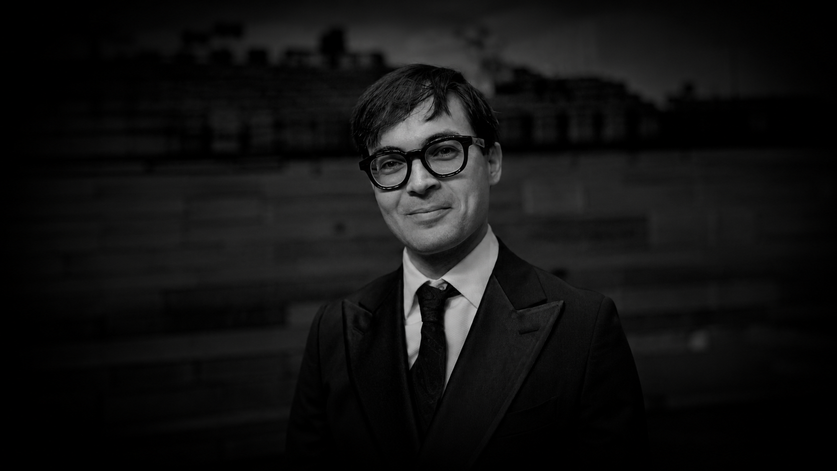 Black and white photo of a man dressed in a suit and tie, wearing glasses, smiling in front of a textured wall.