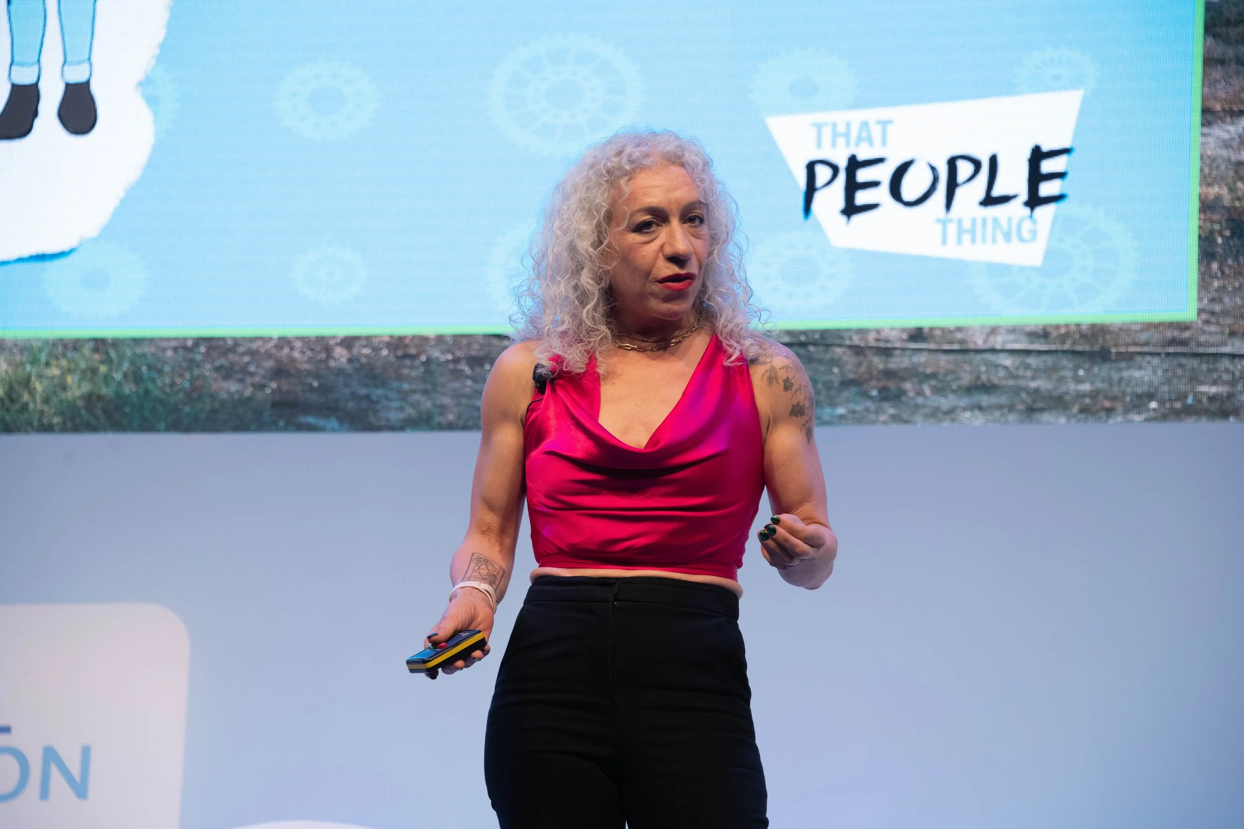 A woman with curly gray hair wearing a pink sleeveless top and black pants, speaking at a presentation or conference, holding a remote control in her right hand, with a background showing a sign that says 'That People Thing'.