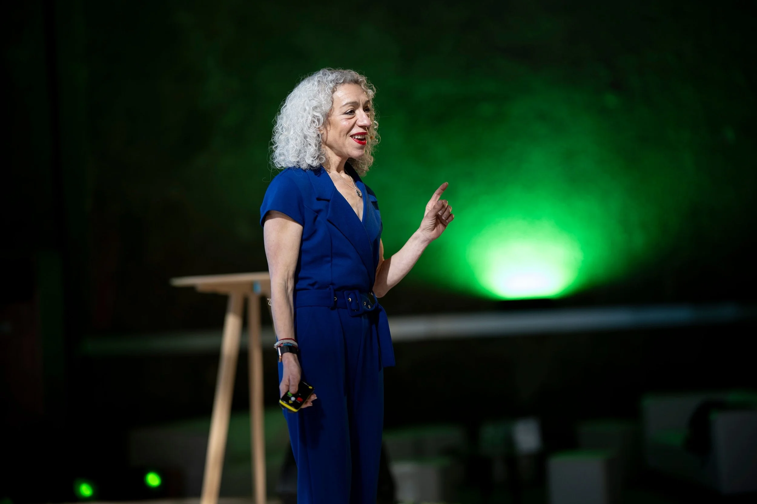 A woman with curly gray hair wearing a blue dress speaking on stage with a green light background.