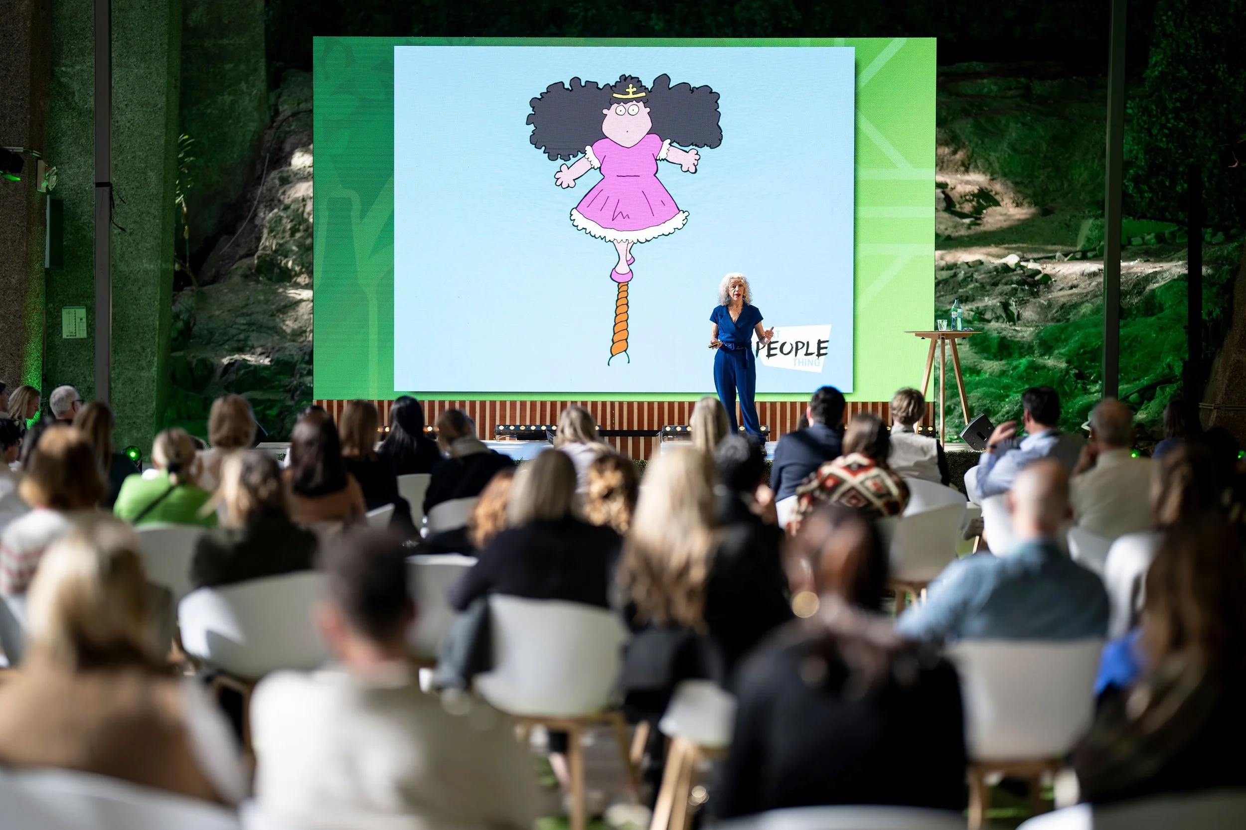 A woman with curly white hair giving a presentation in front of a large screen displaying a cartoon character in a pink dress and a crown. The audience is seated outdoors in a natural setting.
