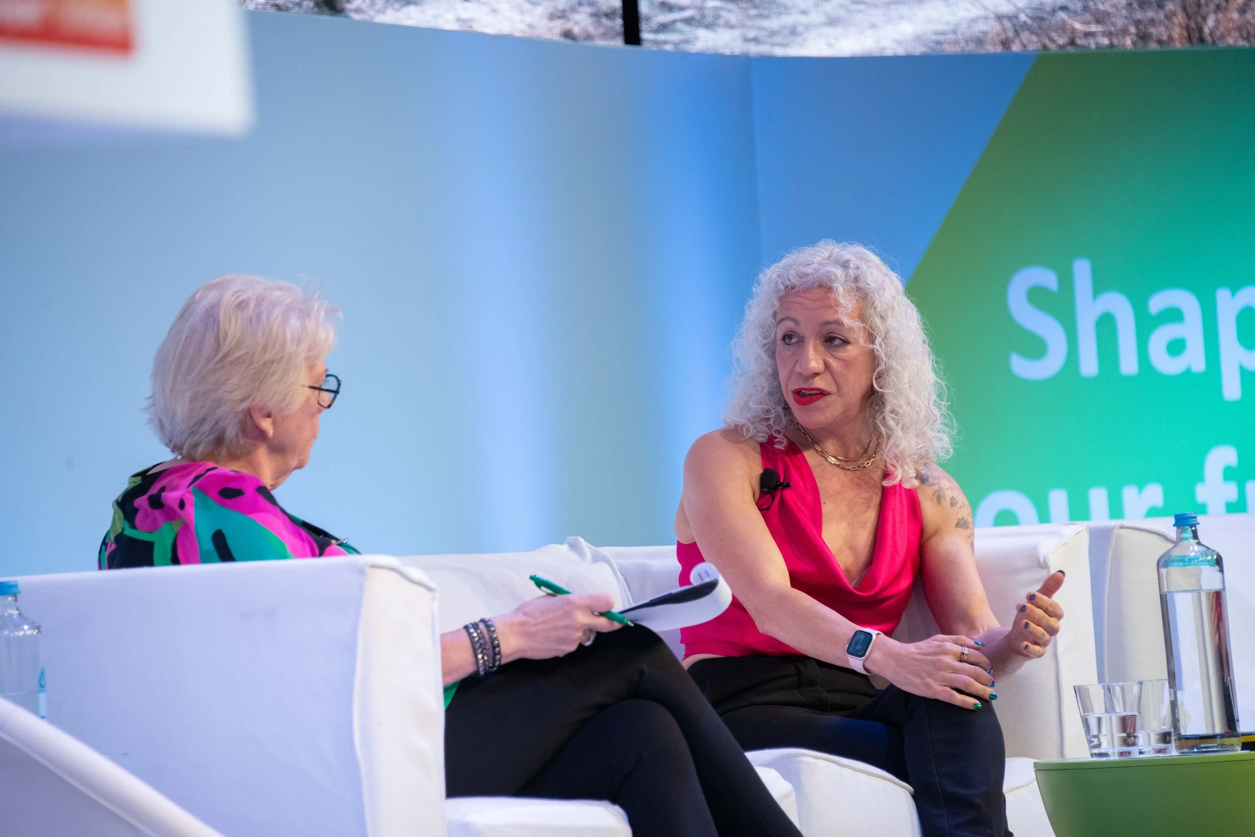 Two women sitting on a stage at a panel discussion. One woman is wearing glasses with curly blonde hair and a colorful dress, and the other woman has curly gray hair, tattoos on her arms, and is wearing a bright pink top.