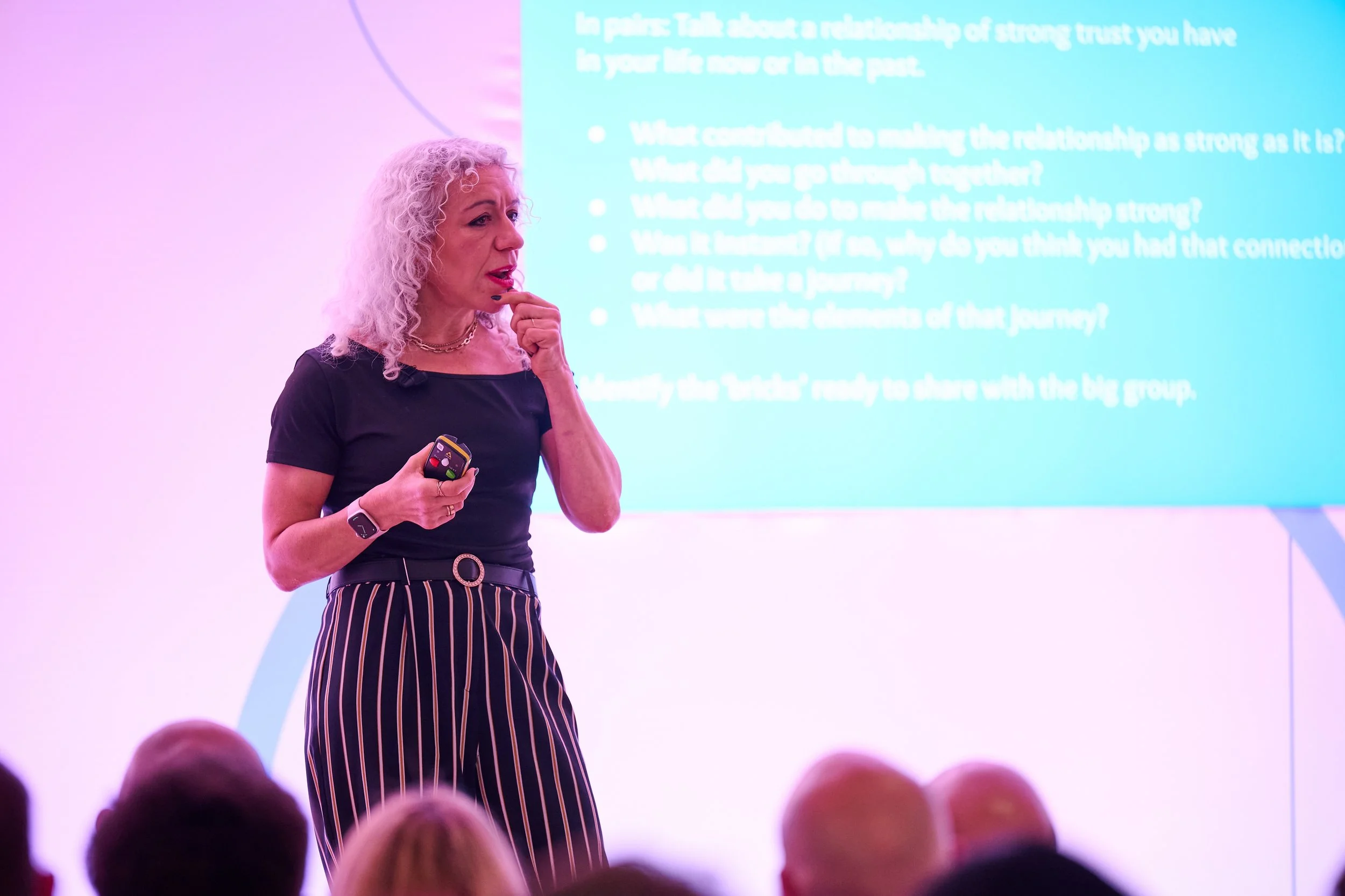 A woman with curly white hair giving a presentation in front of a blue and purple background with a large screen displaying text. She is holding a remote control and appears to be speaking to an audience.