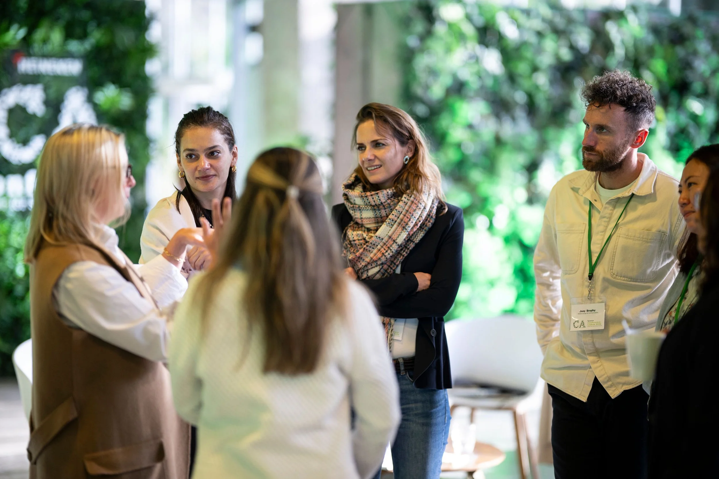 A group of diverse people engaged in conversation at a networking event or conference, with a green, tree-filled background.