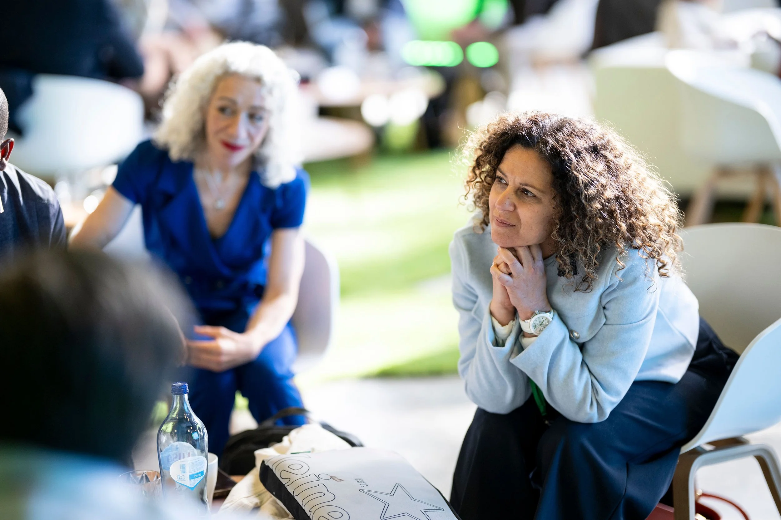 Two women engaged in conversation; one with curly hair and thoughtful expression, the other with gray hair and a blue shirt, sitting at a table outdoors.