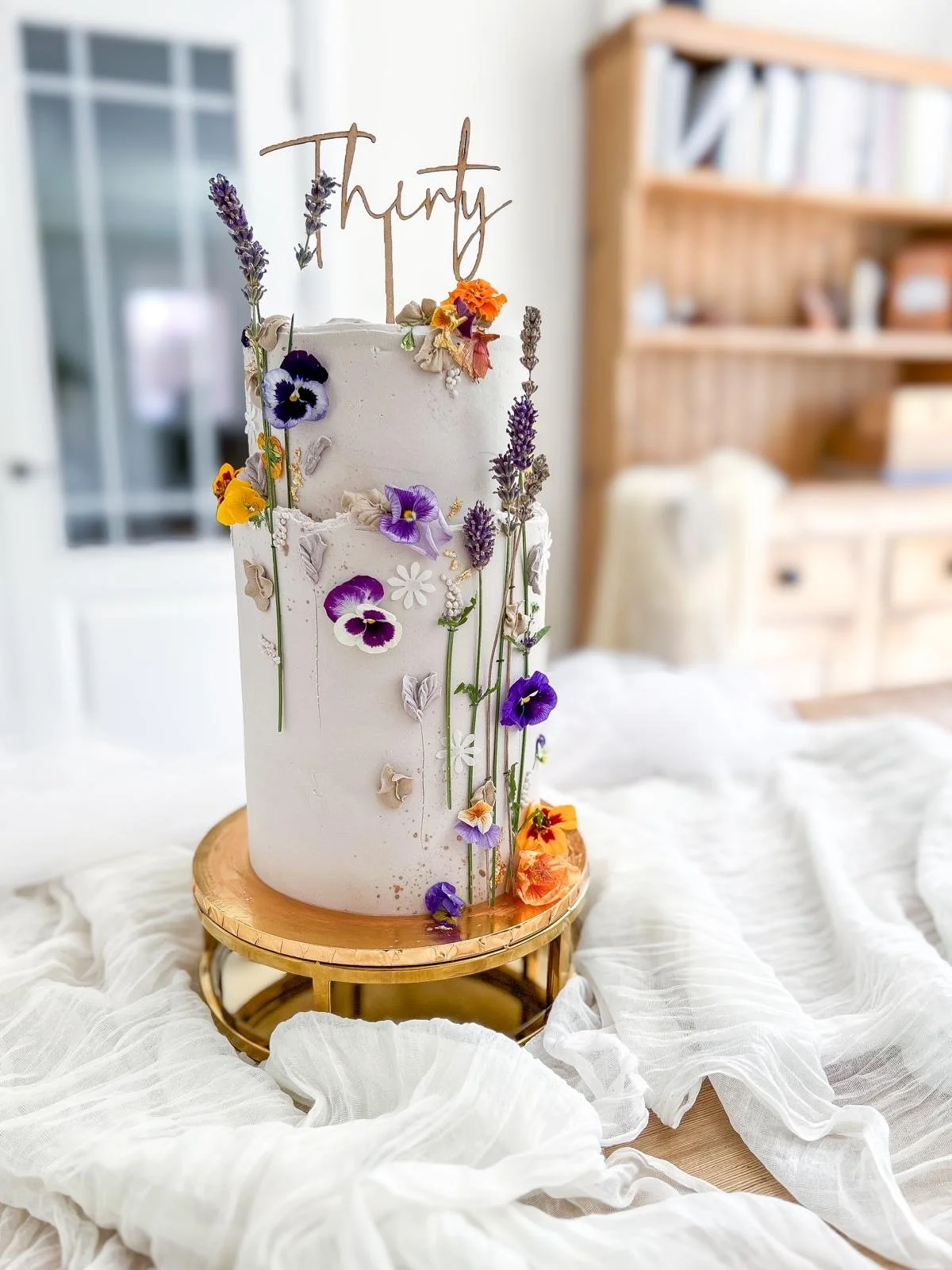 A tall, white, multi-tiered birthday cake decorated with purple, orange, yellow, and cream flowers and greenery, topped with a wooden sculpture spelling "Thirty" in cursive. The cake is on a round, gold stand on a white, crinkled cloth, with a background of a room with a wooden bookshelf and a window.