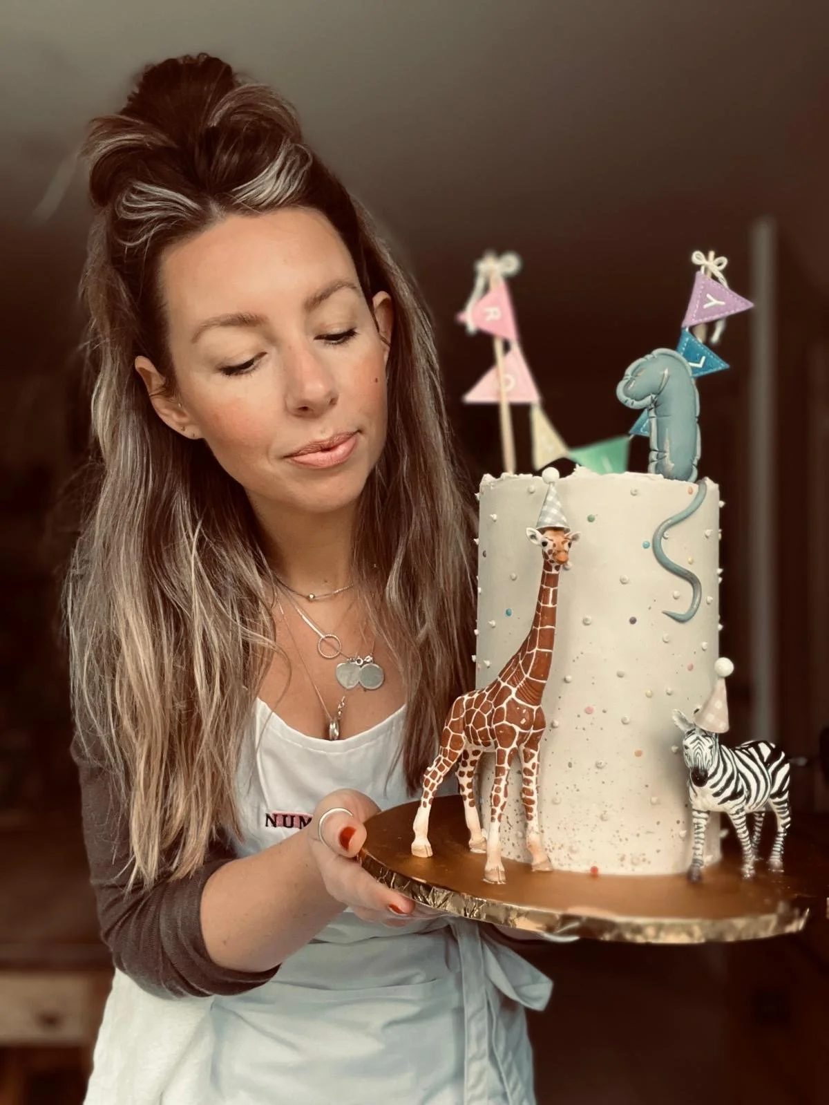 A woman holding a birthday cake decorated with animal figurines including a giraffe, zebra, and elephant, and colorful bunting on top.