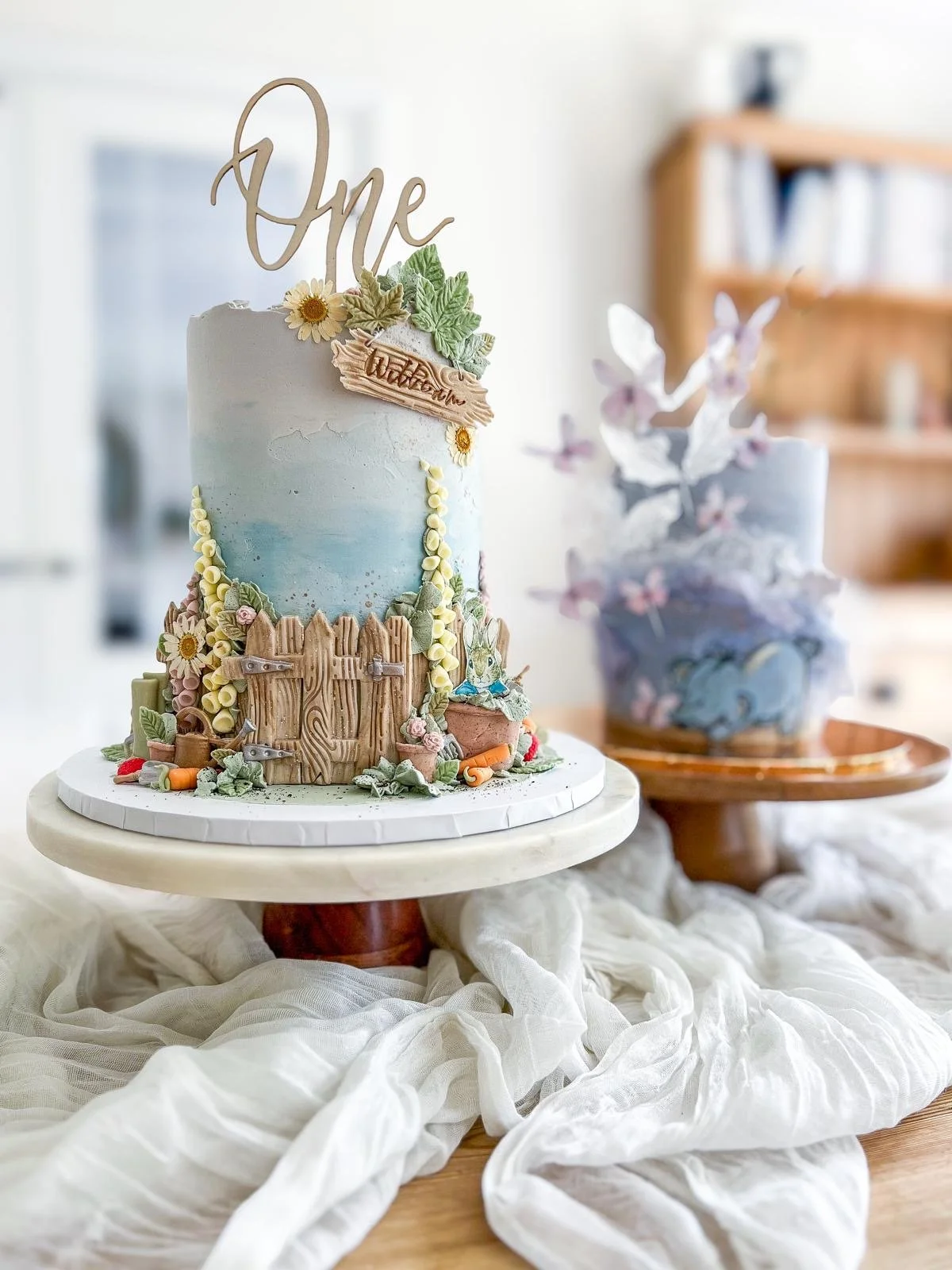 A decorated celebration cake with a woodland theme featuring a wooden fence, bunny figure, flowers, and a topper that says 'One', placed on a white cake stand on a table draped with white fabric.