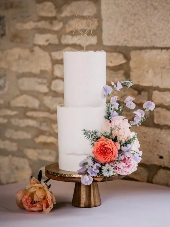 Two-tier white cake decorated with pastel flowers on a wooden cake stand against a stone wall.