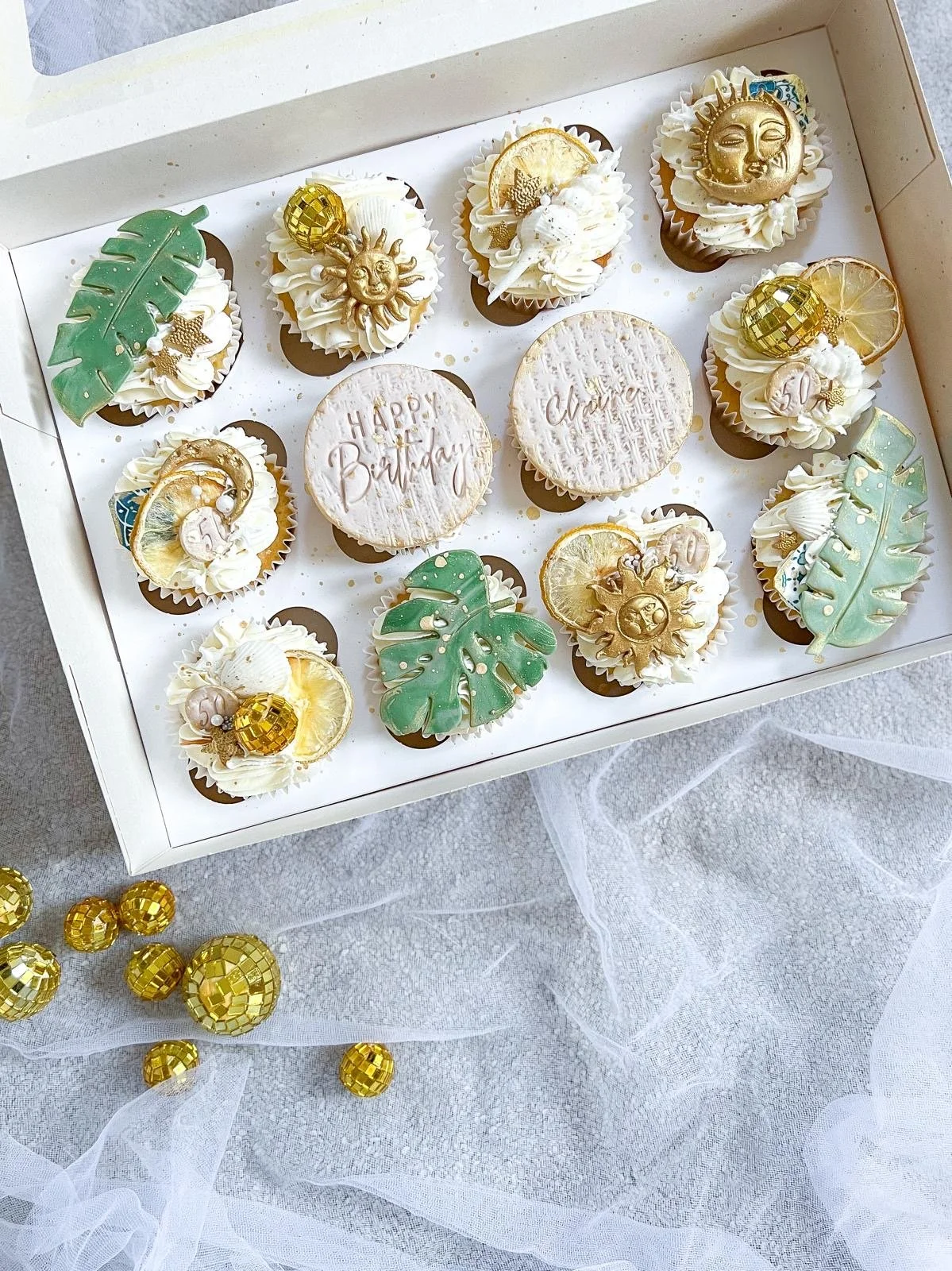 A white box containing ten decorated cupcakes with gold, white, and green tropical and celestial themed decorations, including leaves, sun faces, moon faces, and citrus slices. Two large cookies with engraved messages are in the center, one saying "HAPPY Birthday" and the other "Chase." Gold foil balls and decorative elements are also present around the box and on a sheer white fabric tablecloth.